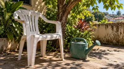 Cadeira e regador de plástico expostos ao sol em um quintal com sombra de folhas